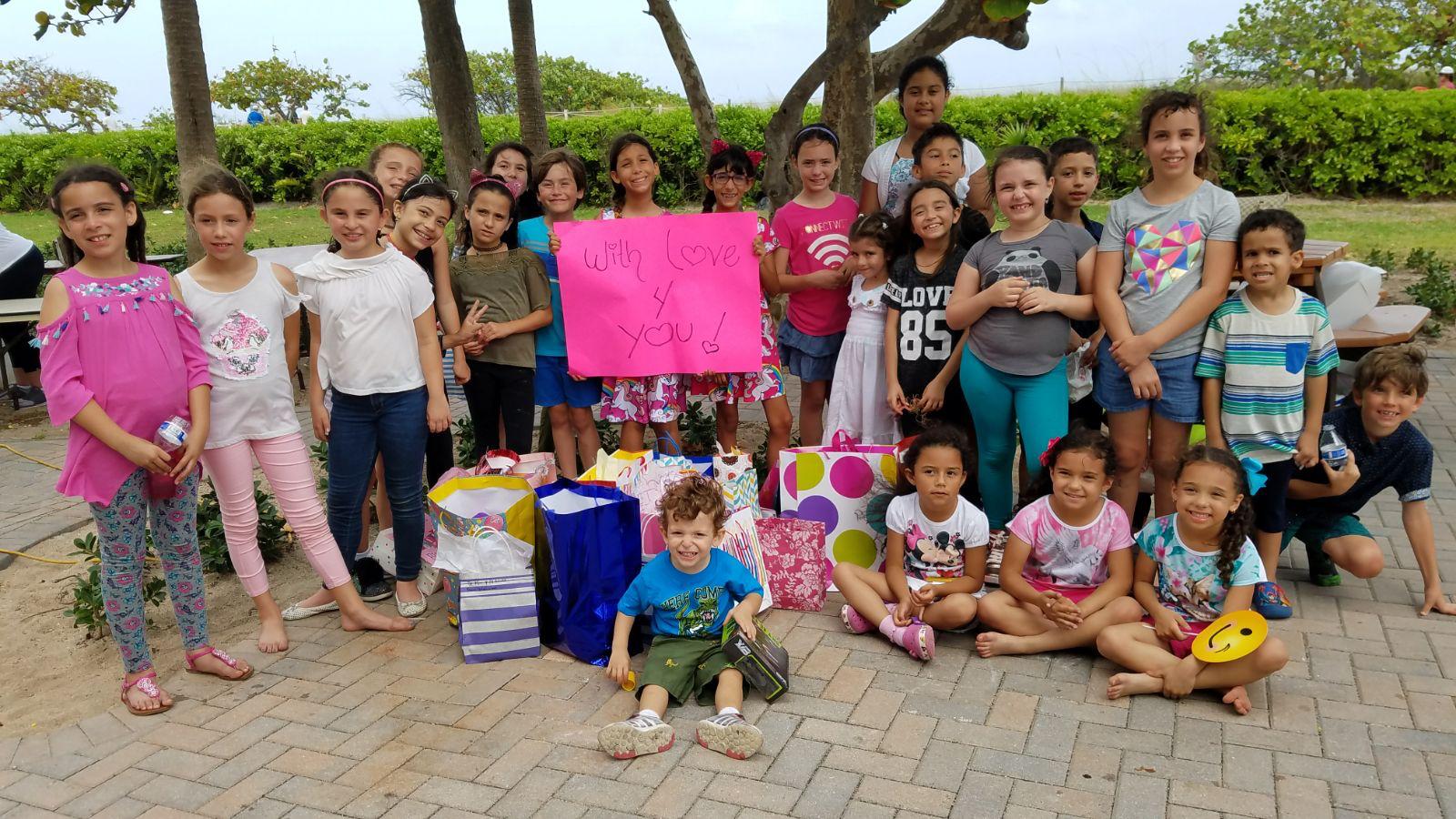 Group of children at the beach holding a handmade 'With Love 4 You' sign next to birthday gift bags